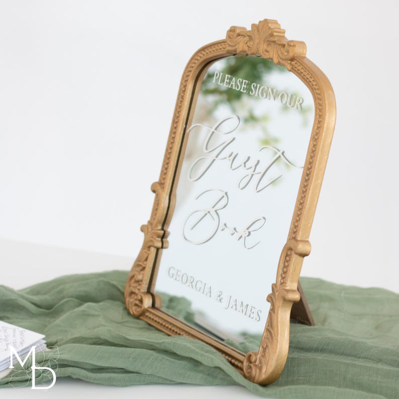 A gold wedding mirror being used as decoration for a wedding entrance table that has a polaroid guest book on it, with the wedding mirror having custom vinyl decal words on it that prompt wedding guests to leave a polaroid and a message as an alternative wedding guest book ideas.