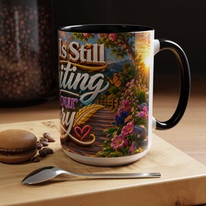 May include: White ceramic mug with a black handle and rim, featuring the text "Is Still Writing Your Story" in gold and teal. The mug's design includes a scenic illustration of a path, flowers, and a butterfly.