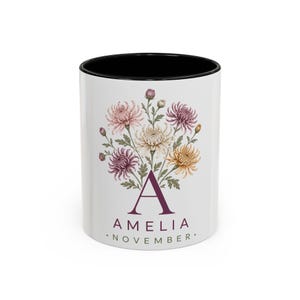 Peut inclure: Un mug en céramique blanche avec un intérieur et un bord noirs, orné d'une illustration florale de chrysanthèmes dans des tons rose, violet et jaune. Sous les fleurs se trouvent une grande lettre A, le nom AMELIA et le mot NOVEMBER en foncé.