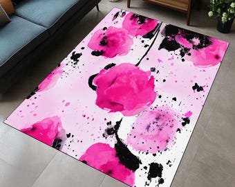 Op de afbeelding: Een rechthoekig vloerkleed met een bloemenontwerp. Grote, felroze bloemen in aquarel-stijl met zwarte accenten zijn verspreid over een witte achtergrond. Roze verfspatten dragen bij aan de artistieke uitstraling. Het vloerkleed ligt op een grijze vloer.