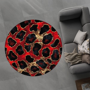 May include: A round rug with a bold animal print design. The rug features a red background with black and gold-colored irregular shapes. The rug is placed on a gray tiled floor, with a gray sofa visible in the background.