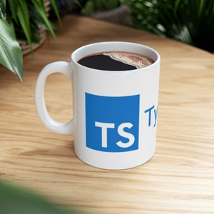 May include: White ceramic mug with a blue square featuring the letters "TS" in white. The mug is filled with black coffee and is sitting on a wooden surface.
