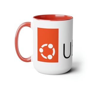 Könnte beinhalten: Eine weiße Keramiktasse mit rotem Rand und Henkel. Die Tasse zeigt ein rotes Quadrat mit einem weißen stilisierten Logo aus drei verbundenen Kreisen. Der Text "Ubuntu" ist in Schwarz unter dem Logo gedruckt.
