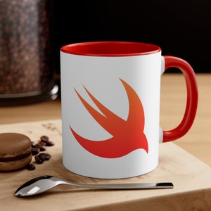 Könnte beinhalten: Weiße Keramiktasse mit rotem Rand und Henkel. Die Tasse zeigt ein rotes und oranges stilisiertes Vogellogo, das Swift-Logo, auf der Vorderseite.