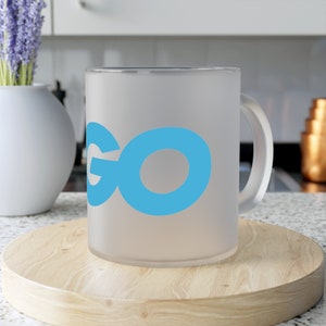 Op de afbeelding: Een matwitte mok met een blauw "GO" logo gedrukt op de voorkant. De mok staat op een houten onderzetter.