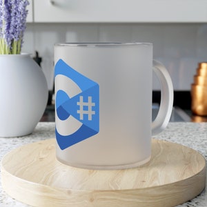 Pode incluir: Uma caneca branca fosca com um logotipo C# azul e branco. A caneca está sobre um descanso de copo de madeira.