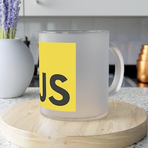 Könnte beinhalten: Eine matte Glas-Tasse mit einem gelben Quadrat und dem Text "JS" in Schwarz. Die Tasse steht auf einem Untersetzer aus Holz.