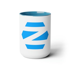 May include: White ceramic mug with a blue interior and a blue geometric design on the exterior. The design is a stylized letter "Z" in a bright blue colour. The mug is cylindrical and has a handle.