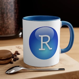 Peut inclure: Une tasse en céramique blanche avec un bord et une poignée bleus. La tasse présente un cercle bleu avec une lettre "R" blanche au centre. La tasse est posée sur une surface en bois avec des grains de café éparpillés autour.