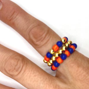 May include: A stack of beaded rings with orange, blue, and gold beads. The rings are worn on a finger.