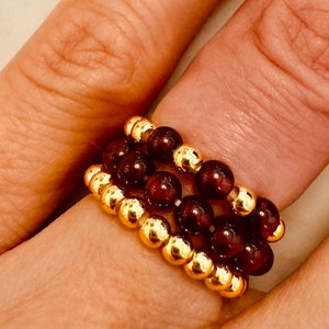 May include: A stack of three rings made of dark red beads and gold beads. The rings are worn on a finger.
