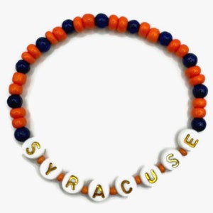 May include: A beaded bracelet featuring the word "SYRACUSE" spelled out in white letter beads with gold lettering. The bracelet has alternating orange and navy blue beads, creating a vibrant color scheme.