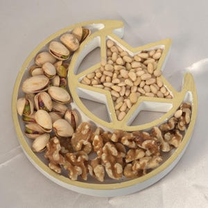Könnte beinhalten: Eine dekorative Servierschale in Form eines Halbmonds und eines Sterns, gefüllt mit verschiedenen Nüssen. Die Schale ist in Abschnitte unterteilt, mit Pistazien, Pinienkernen und Walnüssen. Die Schale hat eine weiße Basis mit goldenem Rand.