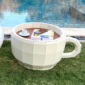 May include: A white, oversized cup and saucer shaped chair and table set. The table has a wooden top and books on it. The set is on a green lawn next to a swimming pool.