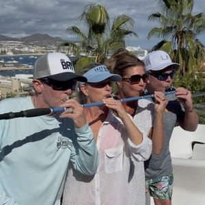 May include: A long, slender, light blue cylindrical tube with black foam grips at both ends, held horizontally by four people. The tube features a series of small, evenly spaced circular openings along its length, designed for simultaneous use.