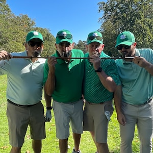 May include: Four men wearing green hats with shamrocks on them are holding a wooden stick in their mouths. They are all wearing green shirts and khaki shorts. They are standing on a green grassy field.