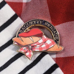 May include: A gold-colored enamel pin featuring a baguette wearing a red beret and a checkered scarf. The pin has the words "BAGUETTE BRIGADE" in a semi-circle around the baguette. The pin is set against a red and white checkered background and a black and white striped shirt.