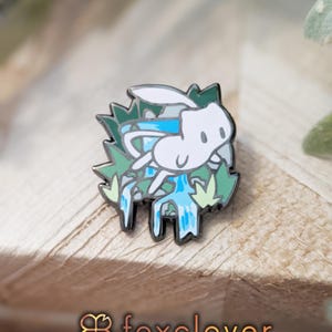May include: A white and blue enamel pin featuring a cartoon creature with green foliage accents. The pin has a silver outline and is set against a wooden surface. The text "foxclover" is at the bottom.