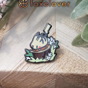 May include: Enamel pin featuring a cartoon character with a brown body, tan face, and black accents, surrounded by green grass and white flowers. The pin has a black outline and is set against a wooden background. The text "foxclover" is in the upper left corner.
