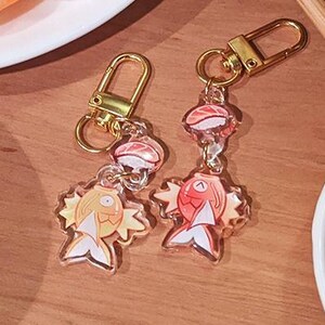 May include: Two keychains featuring a cartoon fish with a sushi nigiri on top. The fish is orange, red, and yellow. The sushi is pink and white. The keychains are on a wooden surface.