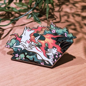 May include: A detailed enamel pin featuring a white and red mythical creature design with gold trim. The pin is set against a backdrop of green foliage and a wooden surface. The brand name "foxclover" is at the bottom.
