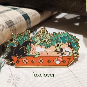 Puede incluir: Un pin rectangular de esmalte con un zorro durmiendo y un gato negro en una jardinera con follaje verde. La jardinera es naranja con ribetes dorados y detalles decorativos. El texto "foxclover" está en la parte inferior.