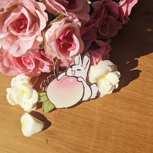 May include: A white enamel pin featuring a cartoon bunny holding a pink peach. The pin is surrounded by pink roses and white flowers.