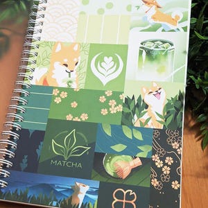 May include: A spiral-bound notebook featuring a patchwork design with green and white squares. The squares display illustrations of Shiba Inu dogs, matcha tea, and floral patterns. The word "MATCHA" is printed on one of the squares. The notebook is on a wooden surface.