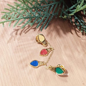 May include: A set of four Christmas light bulb pins in gold, red, blue, green, and yellow. The pins are connected by a delicate gold chain. The background is a light wood surface with a green leafy branch.