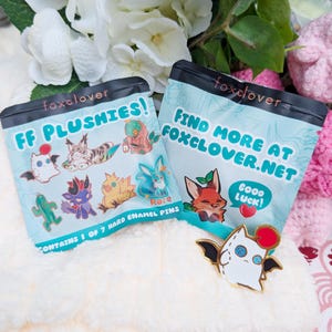 Op de afbeelding: Twee lichtblauwe zakjes met de tekst "FF PLUSHIES!" en "FIND MORE AT FOXCLOVER.NET". De zakjes bevatten harde emaille pins met stripfiguren. Een witte emaille pin met zwarte vleugels en een rode bal is ook zichtbaar.