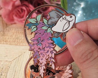 May include: A rose gold metal pin featuring a white and grey fish with a red accent, surrounded by a circle and purple wisteria. The pin also includes green leaves and a second fish design. The pin is held by a hand.