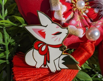 May include: A white enamel pin of a fox with red eyes and a red bow. The fox is sitting on a red tassel.