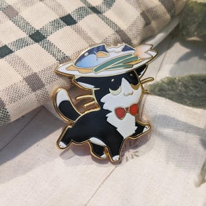 May include: A black and white cat enamel pin with a red bow tie and a hat featuring a fish design. The pin has gold-colored trim and is set against a neutral background.