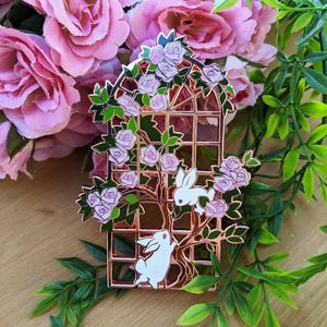 May include: A rose gold metal pin featuring a trellis design with pink roses and two white rabbits. The pin is surrounded by pink roses and green foliage, creating a whimsical and charming aesthetic. This decorative pin is perfect for adding a touch of nature-inspired flair to any outfit.