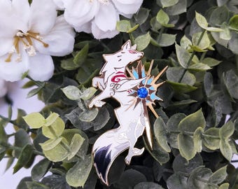 May include: A white and gold enamel pin depicting two stylized foxes with red details and a blue gem. The pin features a gold sunburst design, set against green foliage and white flowers.