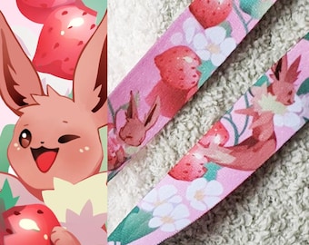 May include: Pink lanyard featuring a cartoon Eevee character winking, surrounded by strawberries. The lanyard also has a repeating pattern of Eevee, strawberries, and white flowers on a pink background. The design is whimsical and colorful.