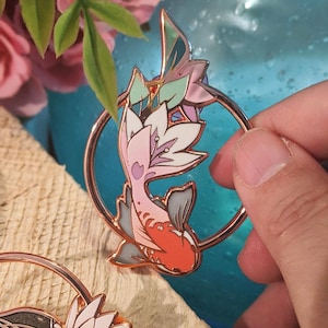 Può includere: Uno spillo in smalto oro rosa con un pesce koi stilizzato con squame rosa, bianche e rosse. Il pesce nuota in un cerchio con un motivo floreale.