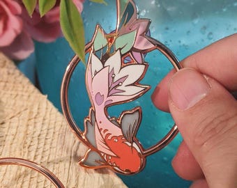 May include: A rose gold enamel pin featuring a stylised koi fish with pink, white and red scales. The fish is swimming in a circle with a floral design.