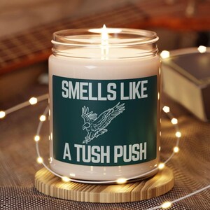 May include: A white candle with a green label that reads "Smells Like A Tush Push". The label also features a stylized image of a bird in flight.