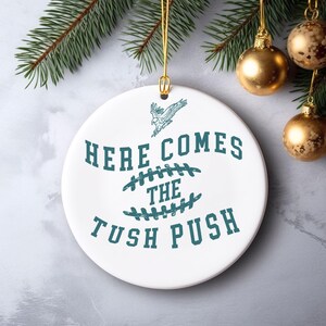 Op de afbeelding: Witte kerstbal van keramiek met een tealgroen ontwerp. De kerstbal heeft een voetbal met stiksels, een vliegende adelaar en de tekst "Here Comes The Tush Push".