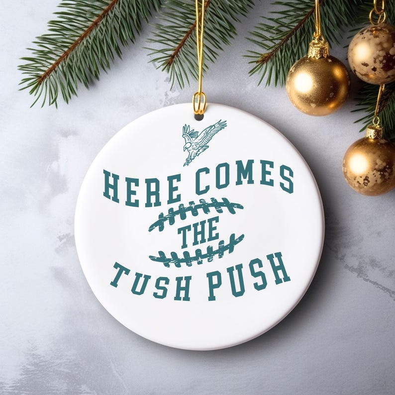 Op de afbeelding: Witte kerstbal van keramiek met een tealgroen ontwerp. De kerstbal heeft een voetbal met stiksels, een vliegende adelaar en de tekst "Here Comes The Tush Push".