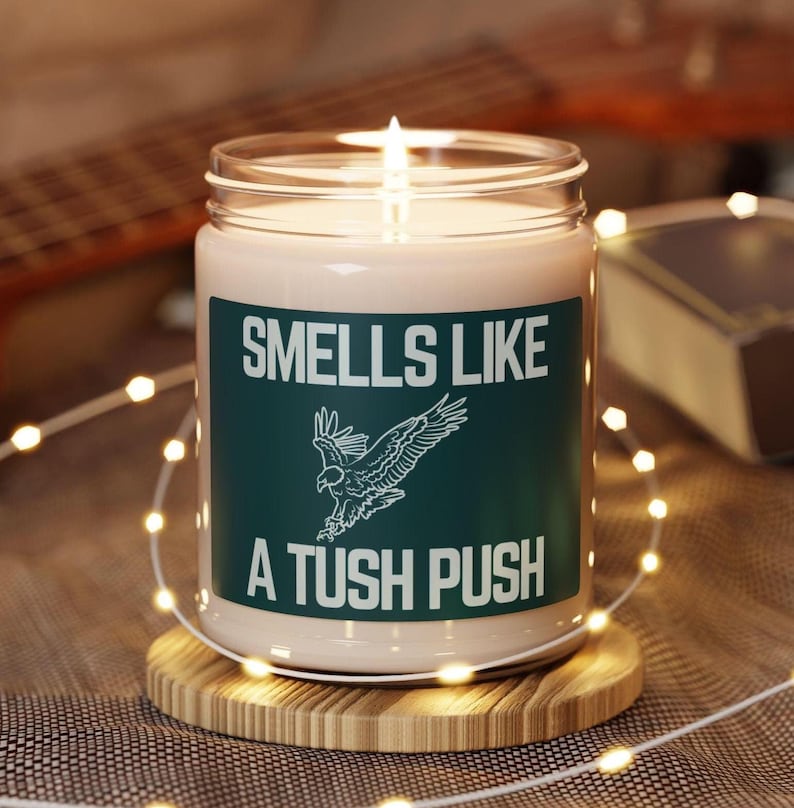 May include: A white candle with a green label that reads "Smells Like A Tush Push". The label also features a stylized image of a bird in flight.