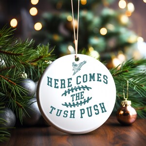 Op de afbeelding: Witte kerstbal van keramiek met een groen ontwerp. De kerstbal heeft een voetbal met stiksels en de tekst "Here Comes The Tush Push".