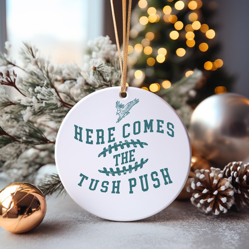 Op de afbeelding: Witte kerstbal van keramiek met een tealgroen ontwerp. De kerstbal heeft een voetbal met de tekst "Here Comes The Tush Push" en een vliegende adelaar.