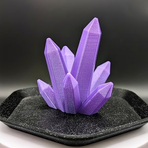 May include: A cluster of purple crystal-shaped objects sits on a black tray. The tray is on top of a white round base.