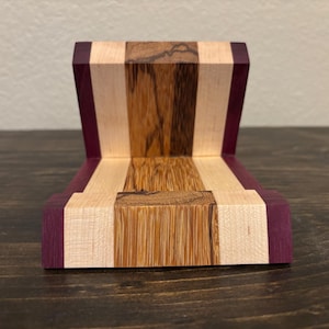 May include: A wooden phone stand with a multi-toned design. The stand features alternating stripes of light wood, dark wood, and purple-hued wood. The stand is designed to hold a phone upright, with a slot at the base for the phone to rest.