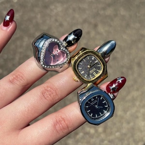 May include: Four unique rings are displayed, each with a distinct design. One is heart-shaped with a pink face, another is gold-toned with a watch face, and two others have watch faces in silver and blue. The rings are held up.