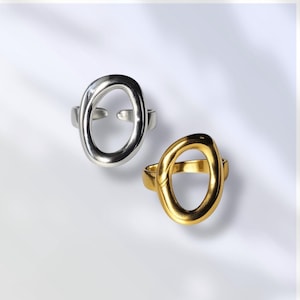 May include: Two open oval rings. One ring is silver-colored, and the other is gold-colored. The rings have a modern, minimalist design. The oval shape is open in the center, creating a unique and stylish look. The rings are set against a white background.