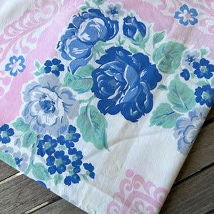 May include: A white fabric textile featuring a floral pattern with large dark blue roses, smaller light blue flowers, and green leaves. The design includes a soft pink decorative border with intricate scrollwork and floral motifs.
