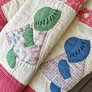 May include: A quilted textile featuring two appliquéd figures with stitched outlines. One figure wears a green bonnet and floral dress, while the other wears a blue bonnet and a checkered dress. The quilt has a red and white patterned border.
